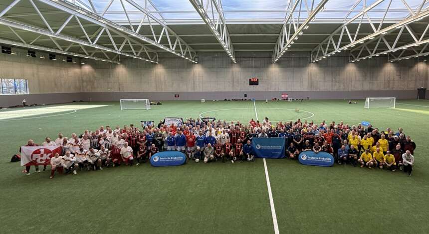 Eine große Gruppe von Menschen, darunter Kinder und Erwachsene, steht auf einem Fußballfeld in einer Sporthalle. Sie sind in verschiedenen Teamfarben gekleidet und posieren für ein Gruppenfoto. Im Vordergrund sind drei Banner mit den Logos der Deutschen Fußball Liga (DFL) und der Deutschen Fußball Akademie (DFA) sichtbar. Die Halle hat ein modernes Design mit einem hohen Dach und großen Fenstern.