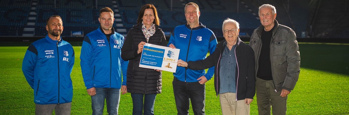 Eine Gruppe von fünf Personen steht auf einem Fußballfeld. Drei von ihnen tragen blaue Jacken mit dem Logo des Vereins und des Sponsors. Eine Frau in der Mitte hält einen Scheck über 1200 Euro in den Händen. Die Szene findet vor leeren Tribünen statt, was auf einen offiziellen Anlass oder eine Spendenübergabe hindeutet.