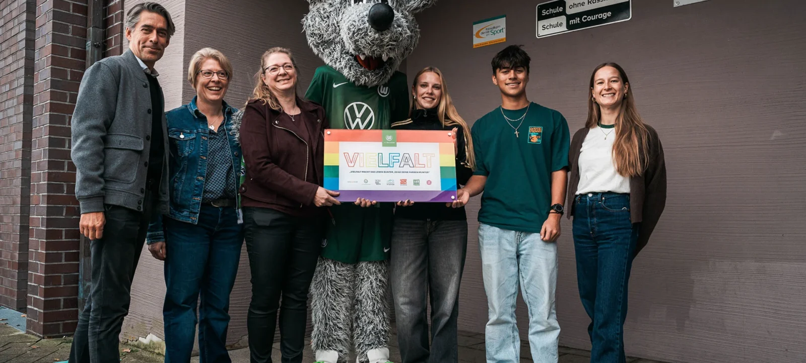 Gruppe von Menschen mit einem Wolf-Maskottchen vor einer Schule, die ein Schild mit der Aufschrift "VIELFALT" halten. Die Schule hat Schilder mit den Aufschriften "Schule ohne Rassismus" und "Schule mit Courage".