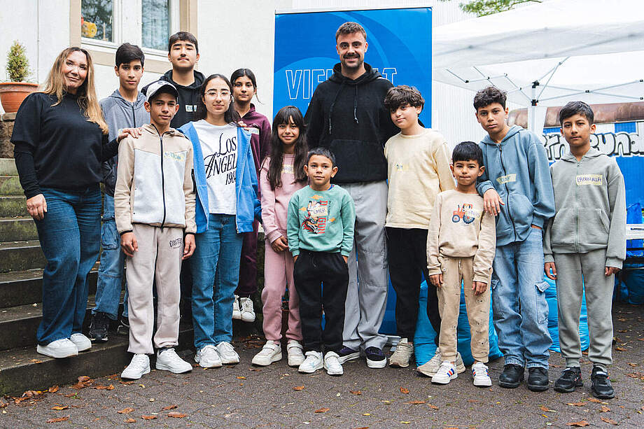 Eine Gruppe von Kindern und zwei Erwachsenen steht vor einem blauen Banner mit der Aufschrift "VIEL". Die Kinder und Erwachsenen lächeln in die Kamera. Die Kinder tragen verschiedene Freizeitkleidung und stehen auf einem gepflasterten Weg vor einem Gebäude mit einem Zelt im Hintergrund.