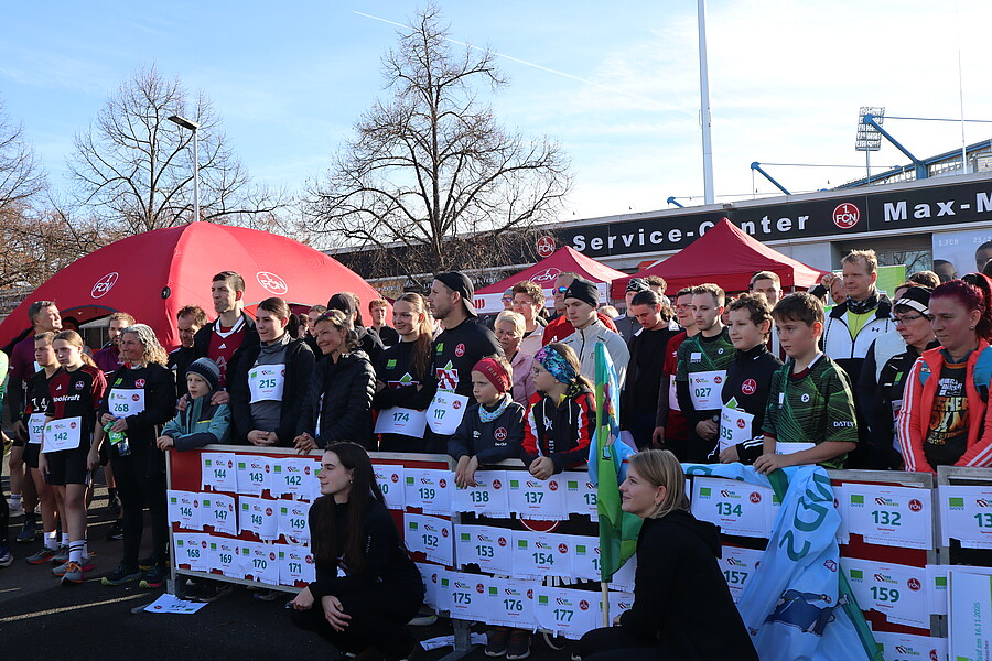 Eine Gruppe von Menschen, darunter Kinder und Erwachsene, steht hinter einem Startblock mit Startnummern. Sie tragen Sportkleidung und sind für einen Lauf oder ein ähnliches Event bereit. Im Hintergrund sind Zelte und ein Service-Center zu sehen, sowie ein Stadion mit kahlen Bäumen und klarem Himmel.