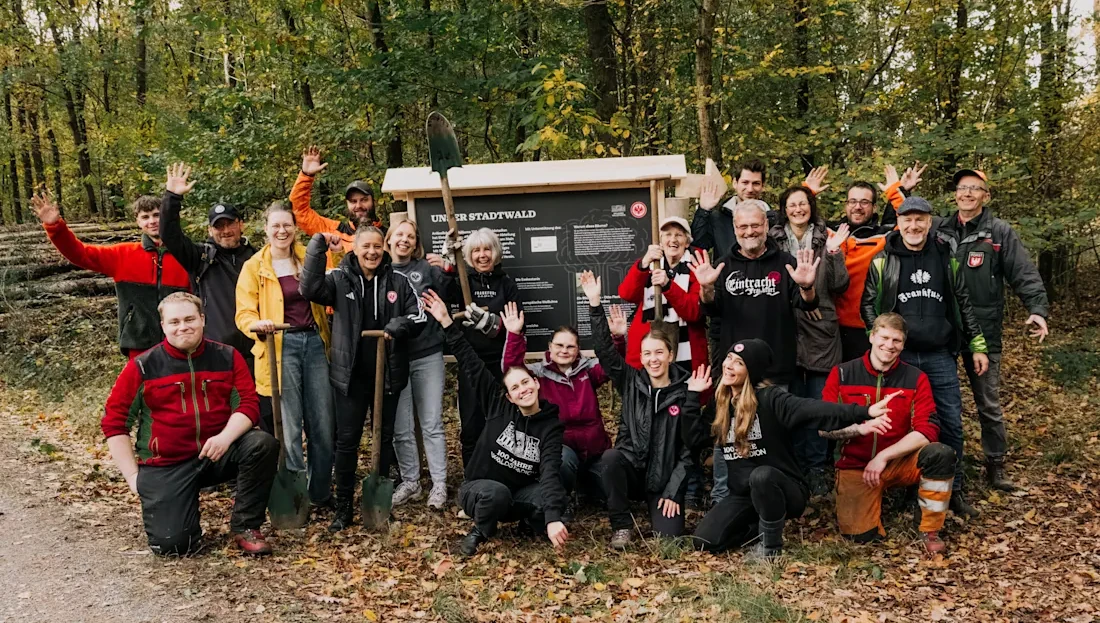 Eine Gruppe von Menschen bei einer Baumpflanzaktion im Frankfurter Stadtwald. Sie stehen vor einem Schild, das "Unter Stadtwald" betitelt ist, und lächeln in die Kamera. Die Teilnehmer tragen verschiedene Jacken und Kleidung, einige halten Schaufeln. Der Hintergrund zeigt einen bewaldeten Bereich mit Bäumen und Laub auf dem Boden.