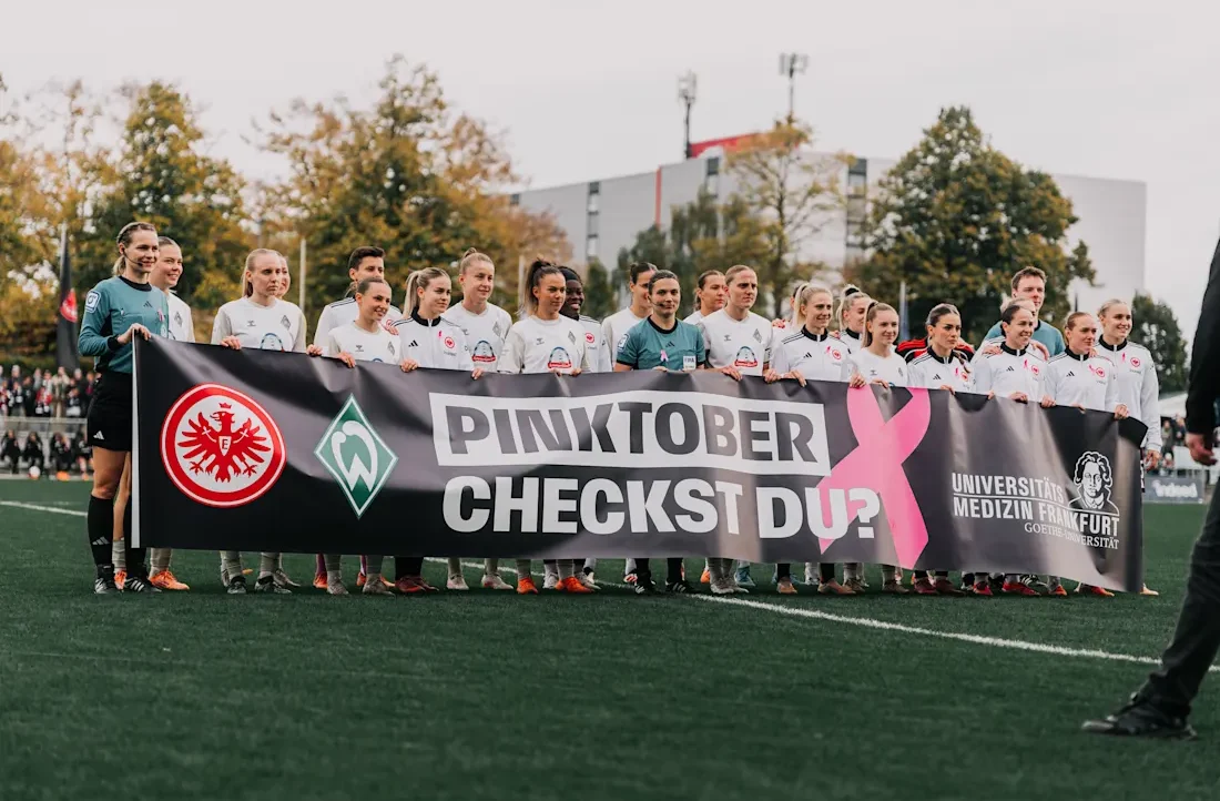 Eine Gruppe von Fußballspielern und Spielerinnen steht auf dem Rasenplatz und hält ein großes Banner. Das Banner zeigt die Logos von Eintracht Frankfurt und SV Werder Bremen sowie den Text "Pinktober Checkst du?" und das Logo der Universitätsmedizin Frankfurt. Die Spieler tragen Trikots mit den jeweiligen Vereinslogos und posieren für ein Gruppenfoto.