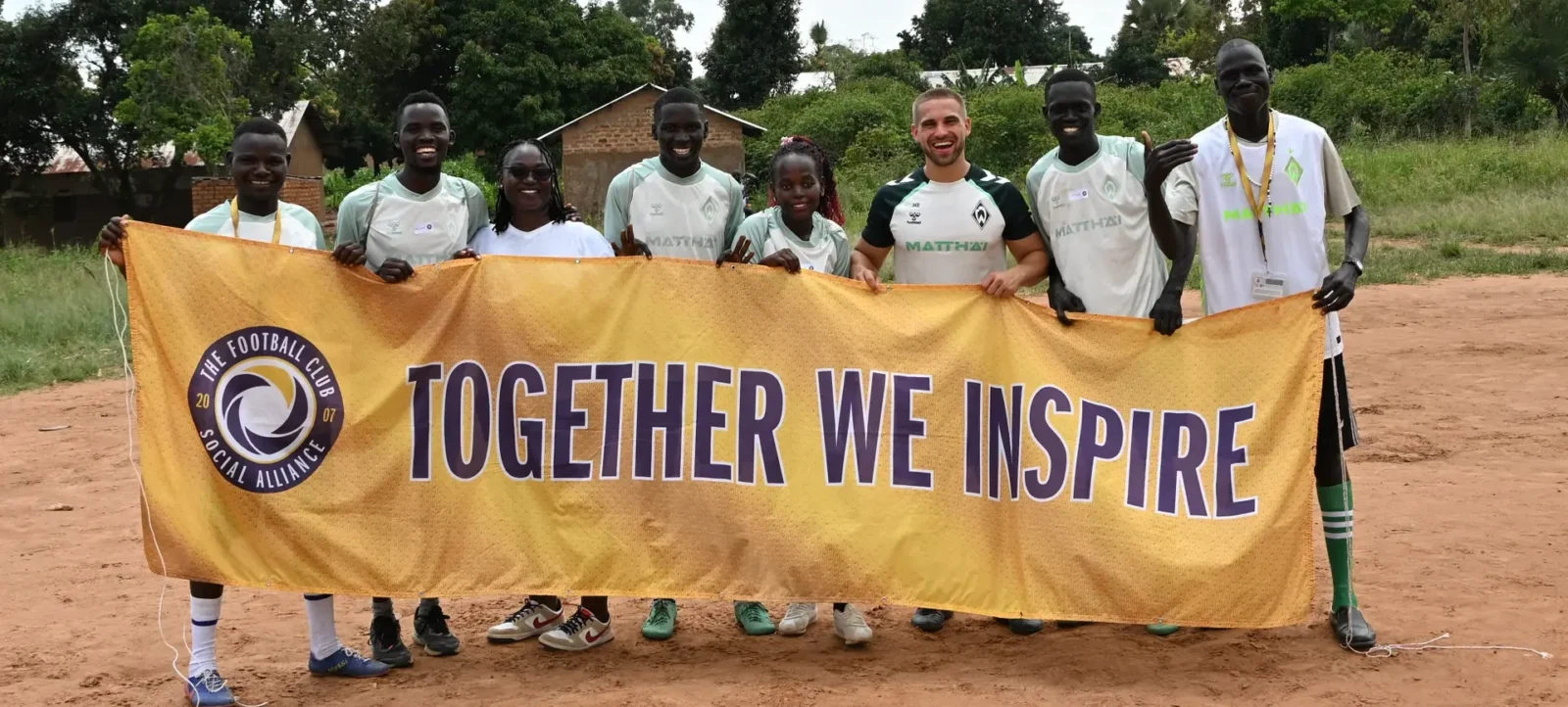 Eine Gruppe von Menschen, darunter Kinder und Erwachsene, hält ein großes gelbes Banner mit der Aufschrift "TOGETHER WE INSPIRE" und dem Logo der "The Football Club Social Alliance 2020". Sie stehen auf einem staubigen Weg vor einer ländlichen Kulisse mit Bäumen und einfachen Gebäuden im Hintergrund. Alle tragen T-Shirts und lächeln in die Kamera.