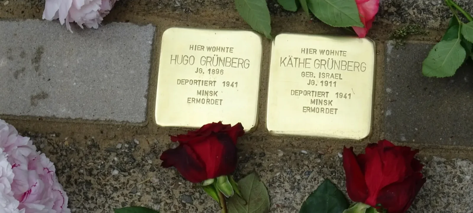 Zwei Stolpersteine auf dem Boden, die an Hugo Grünberg und Käthe Grünberg erinnern. Die Steine tragen die Inschrift, dass sie in Minsk deportiert und ermordet wurden. Um die Steine liegen Blumen, darunter eine rote Rose und eine weiße Blume.