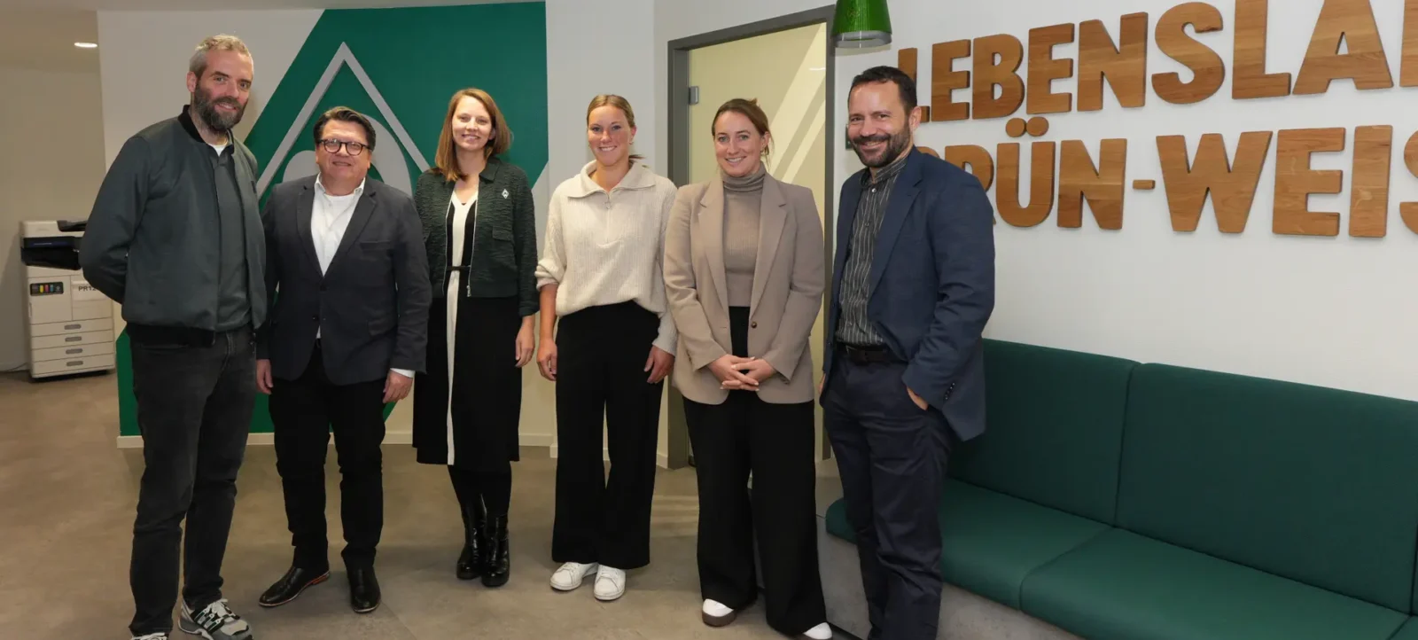 Eine Gruppe von sechs Personen steht in einem modernen Raum mit grüner Wand und einem großen Schild mit der Aufschrift "LEBENSLAU" und "DÜNN-WEI...". Die Personen lächeln und posieren für ein Gruppenfoto. Im Hintergrund ist ein Drucker und eine Tür zu sehen.