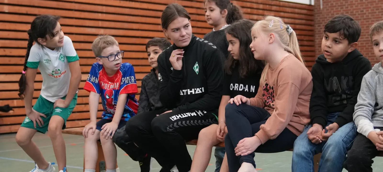 Eine Gruppe von Kindern und zwei Frauen sitzen auf einer Bank in einer Sporthalle. Die Kinder tragen verschiedene T-Shirts und Sportkleidung, einige mit Logos von Fußballvereinen. Die Frauen scheinen in ein Gespräch mit den Kindern vertieft zu sein. Die Halle hat eine Holzvertäfelung und einen grünen Boden.