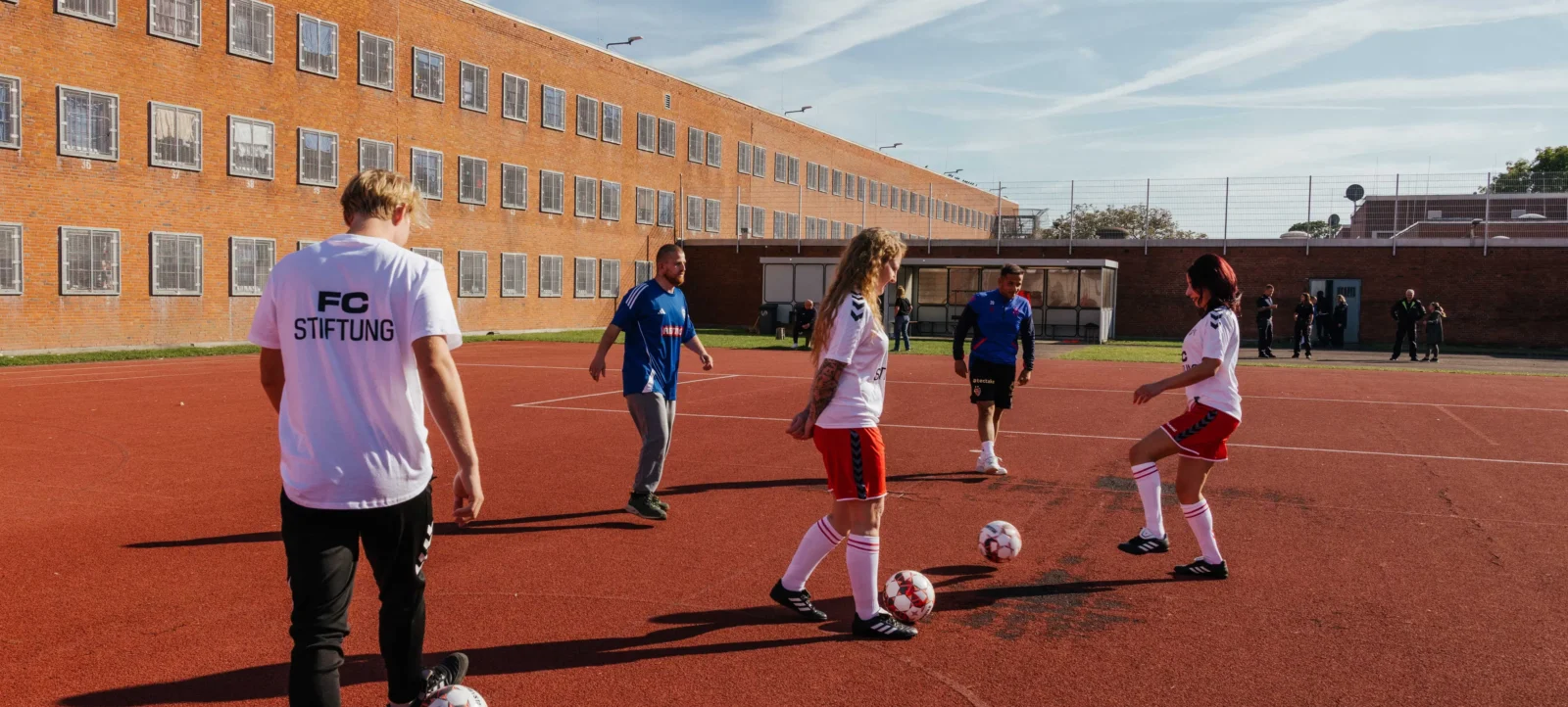 Eine Gruppe von Menschen spielt Fußball auf einem roten Kunstrasenplatz. Im Vordergrund sind zwei Personen zu sehen, die einen Fußball kontrollieren. Eine Person trägt ein weißes T-Shirt mit der Aufschrift "FC STIFTUNG" auf dem Rücken. Im Hintergrund steht ein großes rotes Backsteingebäude mit vielen Fenstern.
