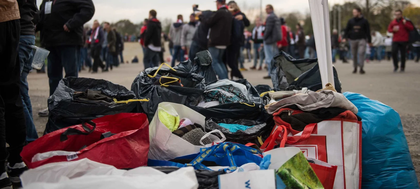 Ein Haufen mit verschiedenen Tüten und Säcken gefüllter Kleidungsspenden, die auf einem Tisch liegen. Im Hintergrund sind Menschen zu sehen, die an einer Spendenaktion teilnehmen. Die Szene findet im Freien statt, möglicherweise auf einem Sportplatz oder einem ähnlichen Gelände.