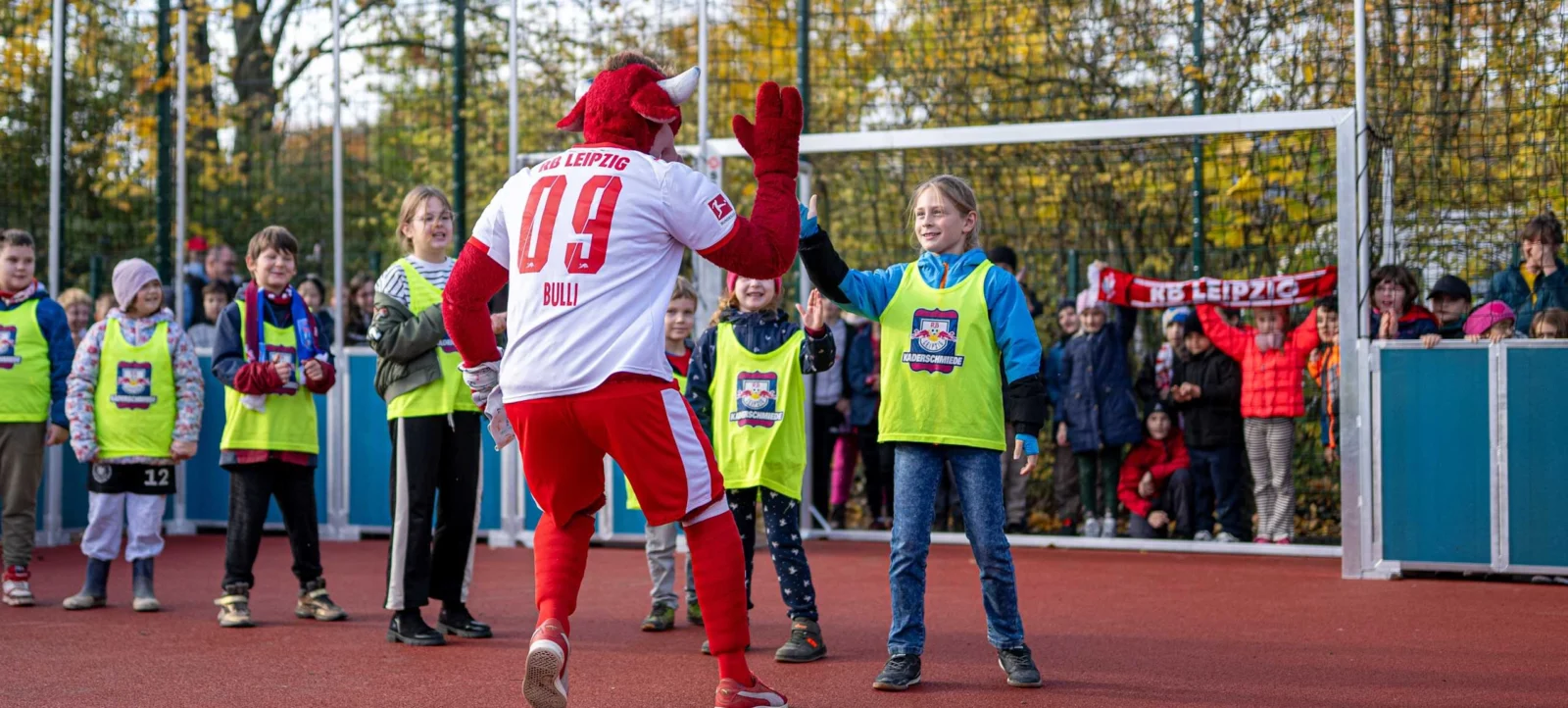 Ein Maskottchen in rot-weißer Kleidung und mit Hörnern auf dem Kopf gibt einem Kind in grüner Weste und blauer Jacke auf einem Sportplatz ein High-Five. Im Hintergrund stehen weitere Kinder und Zuschauer, einige halten eine Schärpe mit dem Aufdruck "RB Leipzig". Die Szene findet auf einem Sportplatz mit rotem Boden und einem Tor im Hintergrund statt.