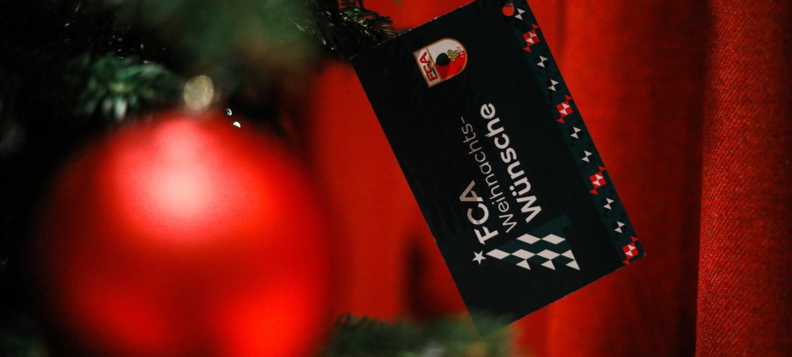 Ein schwarzer Weihnachtsbaumschmuck mit dem Logo des FC Augsburg und der Aufschrift "FC Augsburg Weihnachten" hängt an einem grünen Tannenzweig. Im Hintergrund sind unscharfe rote Lichter und Dekorationen zu sehen.