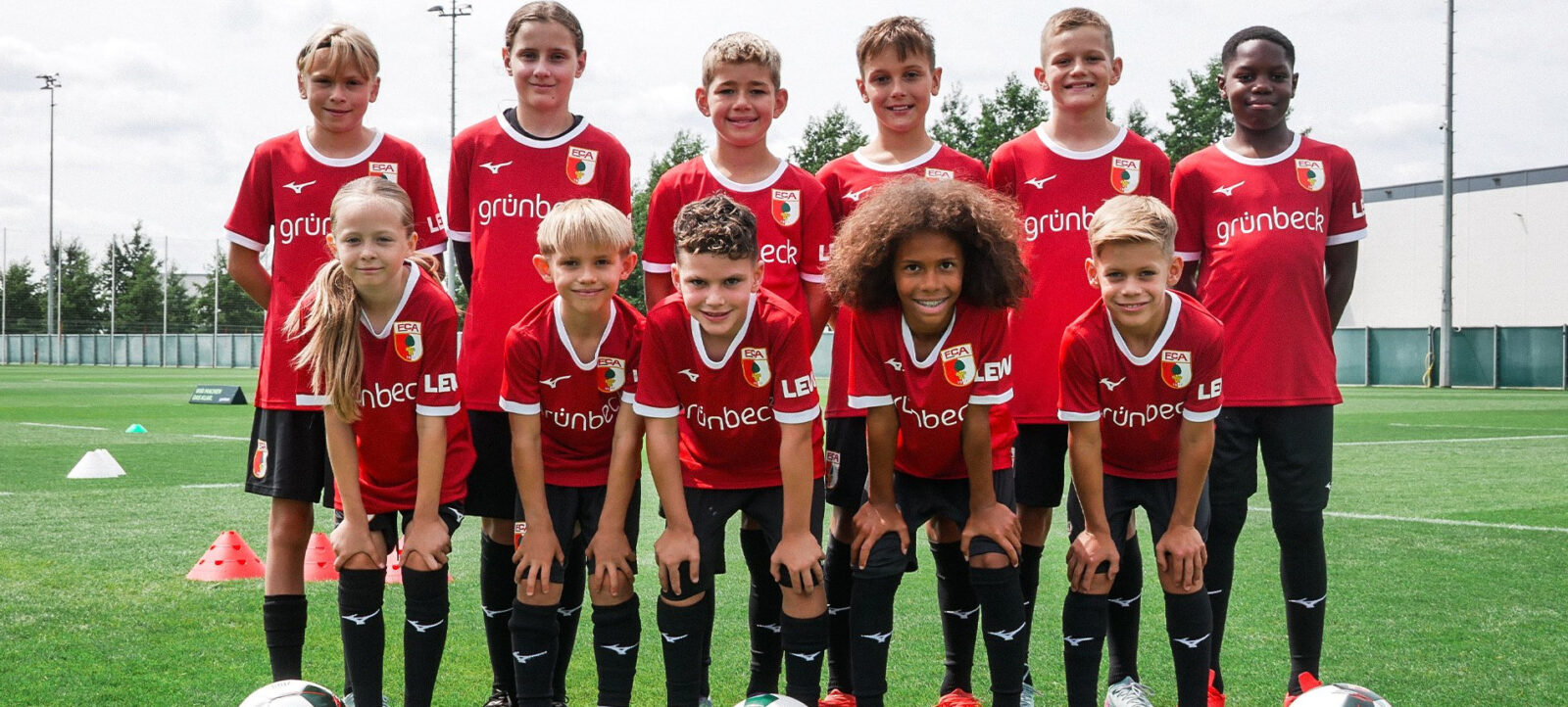 Eine Gruppe von Kindern in roten Fußballtrikots mit dem Aufdruck "grünbeck" steht auf einem Fußballfeld für ein Gruppenfoto. Die Kinder lächeln in die Kamera und posieren mit Fußballschuhen und Bällen vor sich.