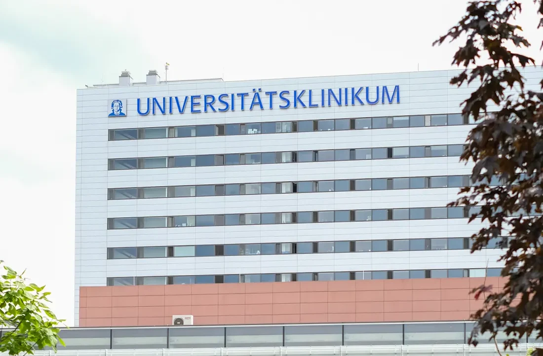 Hochhaus des Universitätsklinikums Frankfurt mit der Aufschrift 'UNIVERSITÄTSKLINIKUM' in blauen Buchstaben auf weißem Hintergrund. Das Gebäude hat mehrere Stockwerke mit Fenstern und ist teilweise von Bäumen verdeckt.