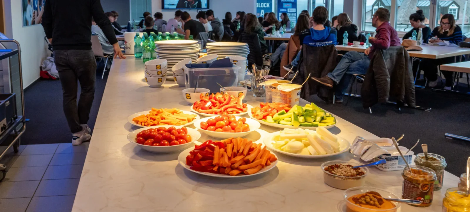 Ein Buffet mit verschiedenen frischen Gemüsesnacks, darunter Paprika, Gurken, Tomaten und Karotten, auf einem Tisch in einem Raum, in dem eine Gruppe von Menschen an Tischen sitzt und arbeitet. Im Hintergrund sind Stapel von Tellern und Getränkeflaschen zu sehen.