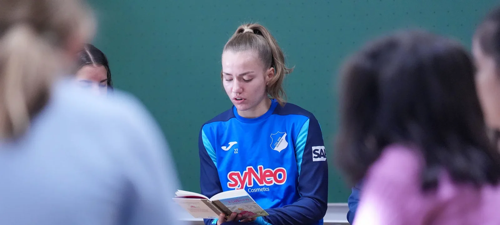 Eine Person in einem blauen Trikot mit dem Logo von syNeo Cosmetics und der Nummer 22 liest konzentriert aus einem Buch vor. Im Hintergrund sind weitere Personen zu sehen, die ebenfalls an der Veranstaltung teilnehmen.