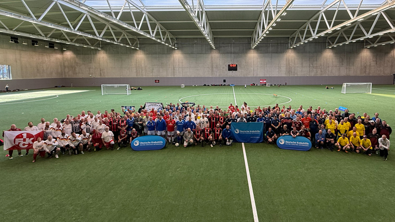 Eine große Gruppe von Menschen steht auf einem Fußballfeld in einer Sporthalle. Die Teilnehmer tragen verschiedene Trikots und sind in mehreren Reihen aufgestellt. Im Vordergrund halten einige Personen Banner und Schilder mit dem Logo "Deutsche Kinder- und Jugendstiftung". Die Halle hat ein modernes Dach mit Stahlträgern und ist gut beleuchtet.