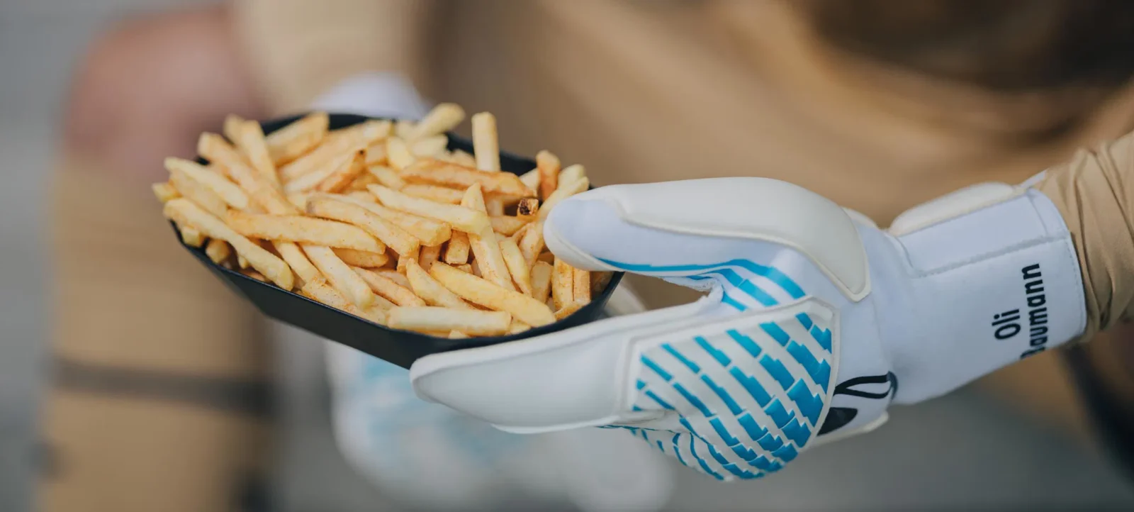 Eine Hand in einem Torwart-Handschuh hält eine schwarze Schale mit Pommes Frites. Der Handschuh ist weiß mit blauen Mustern und trägt den Namen "Oli Baumann". Die Person trägt auch eine braune Handschuhunterlage.