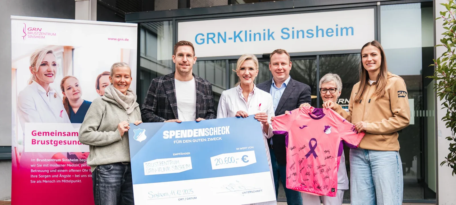 Eine Gruppe von Menschen steht vor der GRN-Klinik Sinsheim und hält einen großen Scheck über 20.600 Euro für das Brustzentrum Sinsheim. Eine Person hält zusätzlich ein signiertes Trikot mit einem pinken Brustkrebs-Awareness-Band. Die Gruppe lächelt und scheint sich über die Spendenübergabe zu freuen.