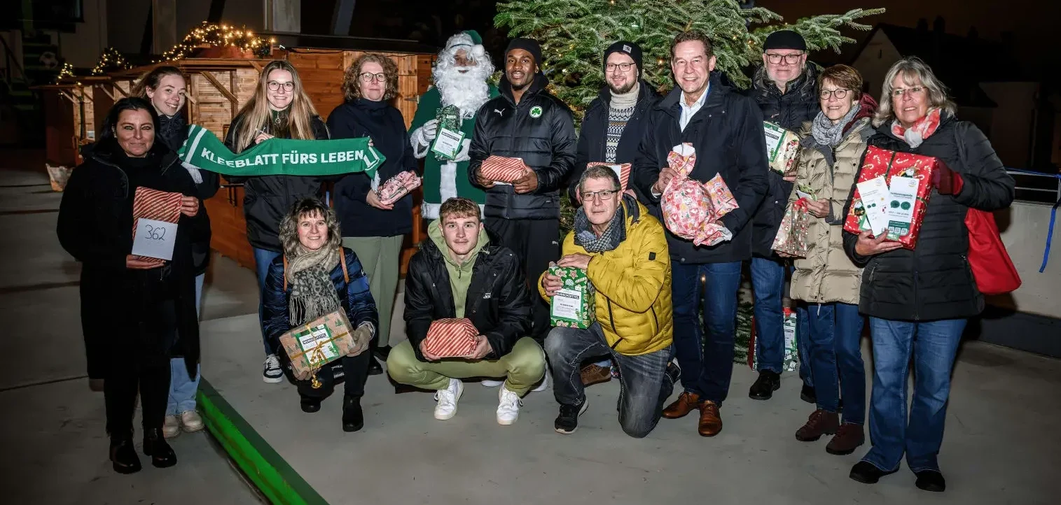 Eine Gruppe von Menschen steht vor einem Weihnachtsbaum und hält Geschenke in den Händen. Eine Person hält ein Schild mit der Aufschrift "KLEEBATT FÜRS LEBEN". Die Gruppe scheint sich an einem Weihnachtsmarkt zu befinden, wie die dekorierten Holzhütten im Hintergrund vermuten lassen.