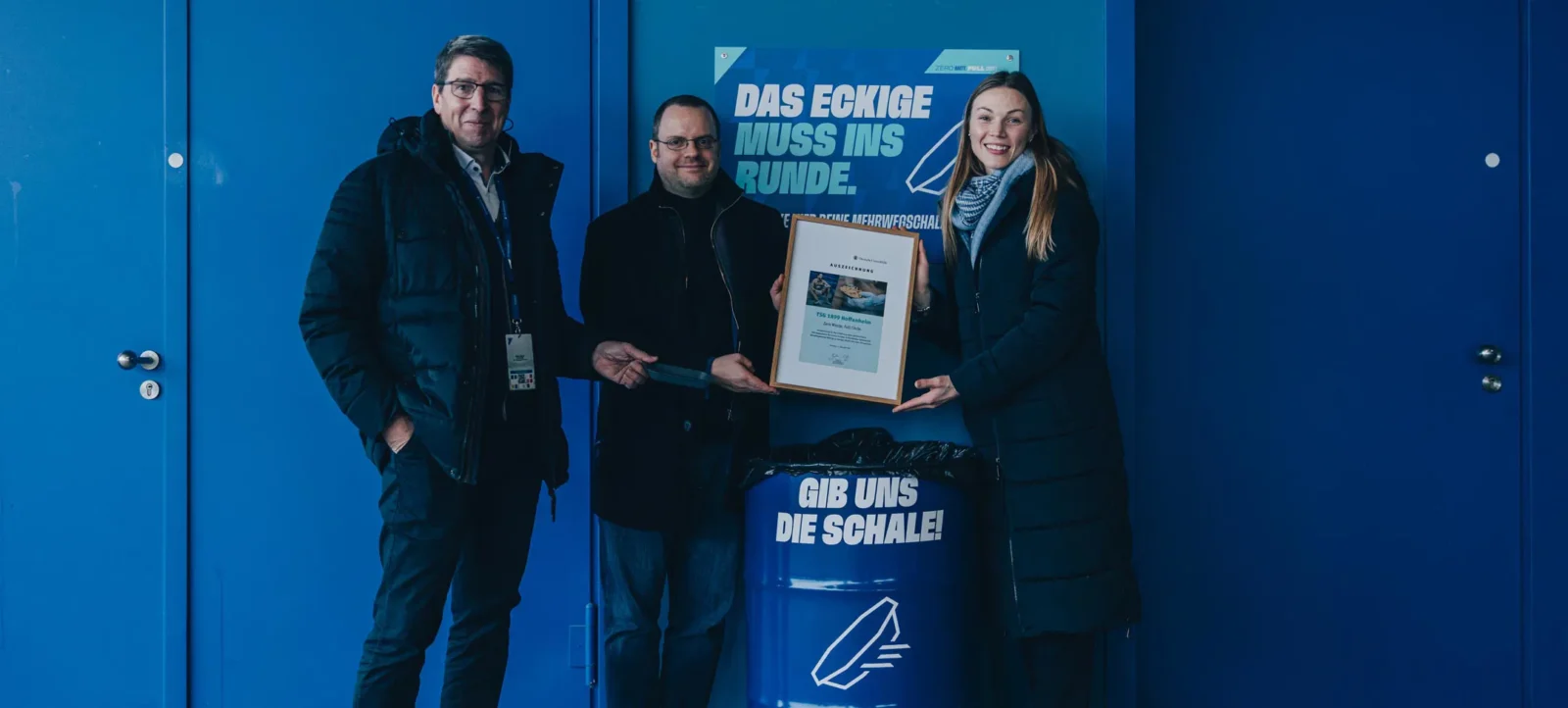 Drei Personen stehen vor einer blauen Tür und halten gemeinsam ein gerahmtes Zertifikat. Eine große blaue Tonne mit der Aufschrift "GIB UNS DIE SCHALE!" und einem Pfeil-Symbol steht vor ihnen. Im Hintergrund ist ein Schild mit der Aufschrift "DAS ECKIGE MUSS INS RUNDE." zu sehen. Die Personen lächeln und scheinen sich über eine erfolgreiche Aktion zu freuen.