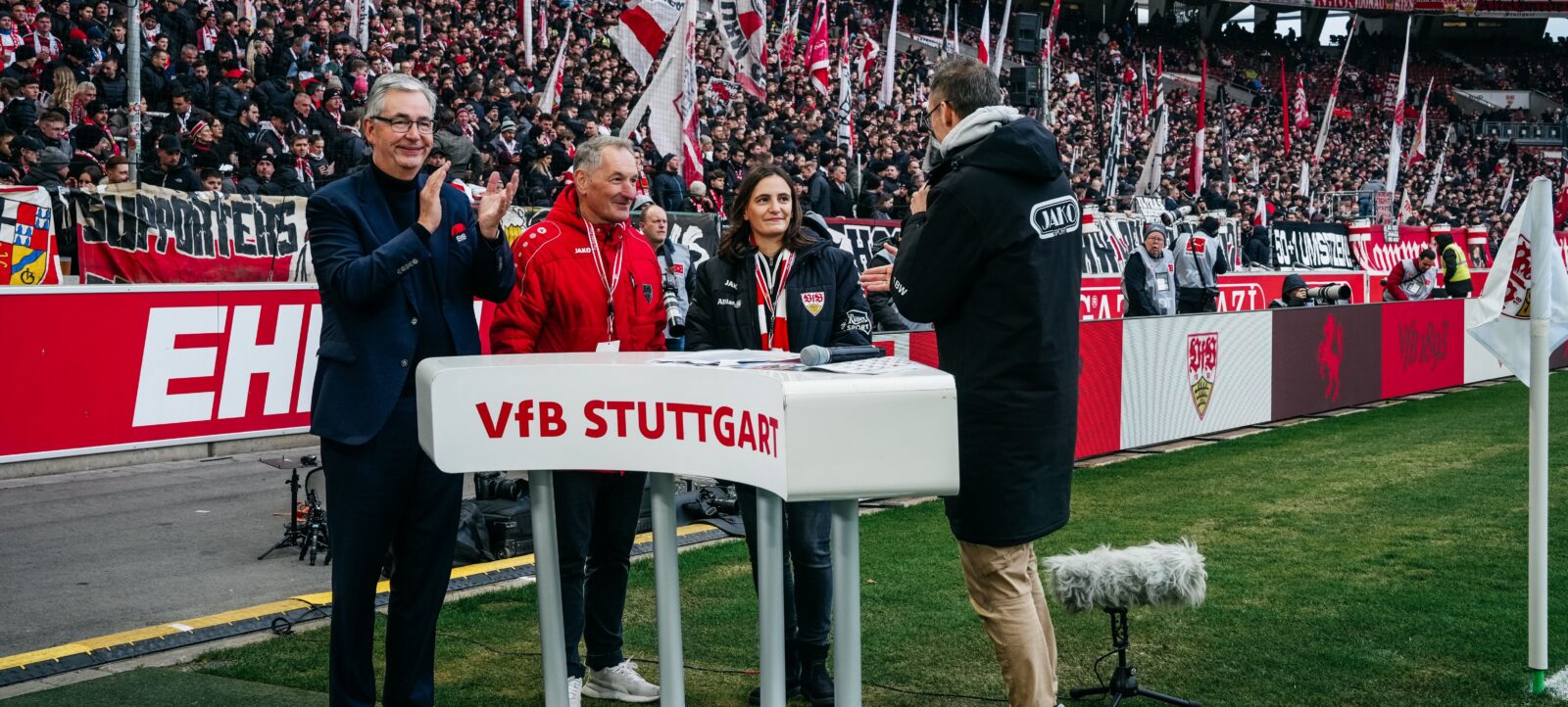Gruppe von Personen am Rednerpult des VfB Stuttgart auf dem Spielfeld, im Hintergrund jubelnde Fans mit Schildern und Fahnen.
