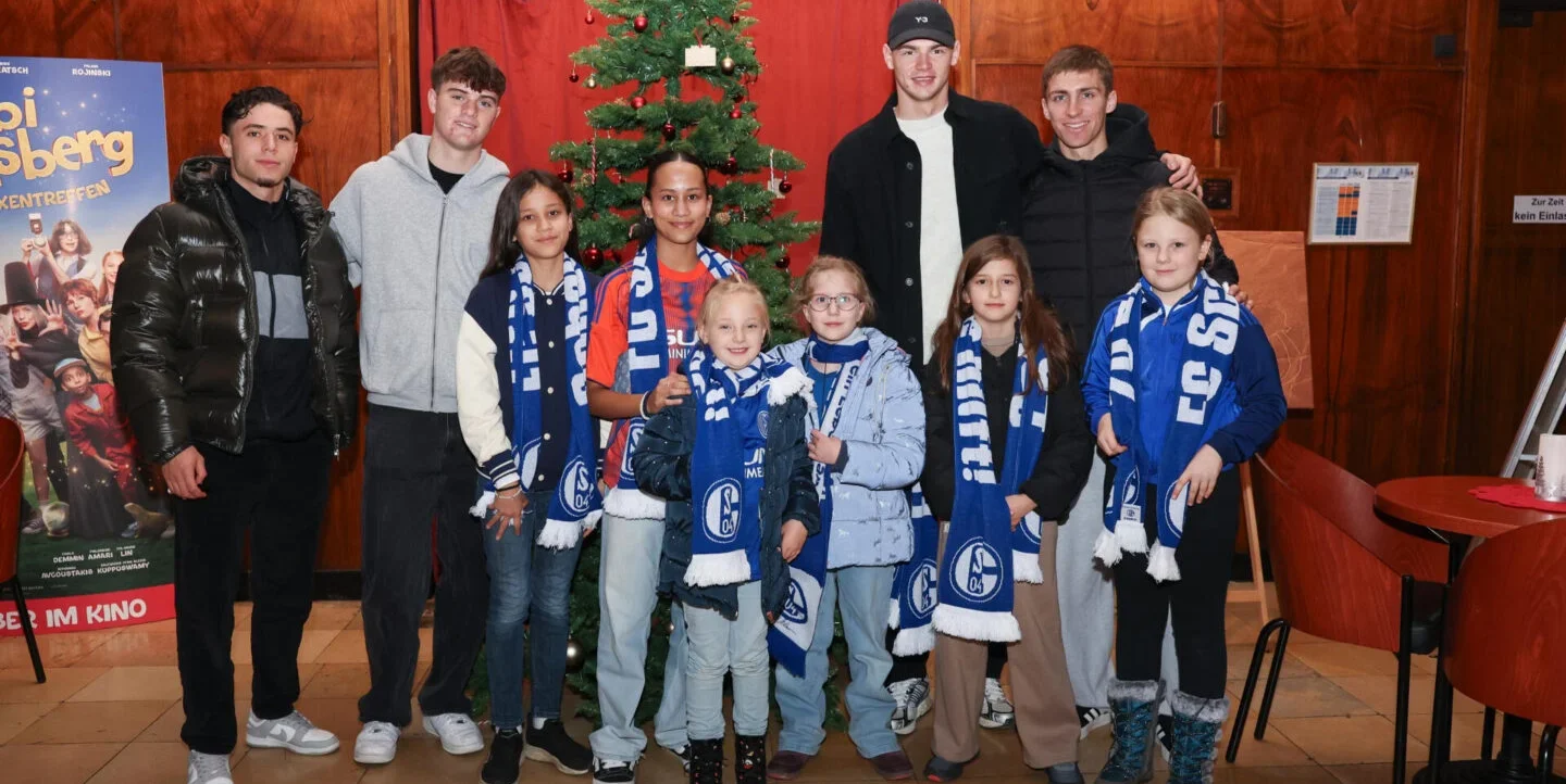 Eine Gruppe von Kindern und Jugendlichen steht vor einem geschmückten Weihnachtsbaum und posiert für ein Gruppenfoto. Sie tragen alle Schals und Hoodies mit dem Logo des FC Schalke 04. Im Hintergrund ist ein Plakat mit dem Titel "Bi Muislberg" zu sehen. Die Kinder und Jugendlichen lächeln und wirken fröhlich.