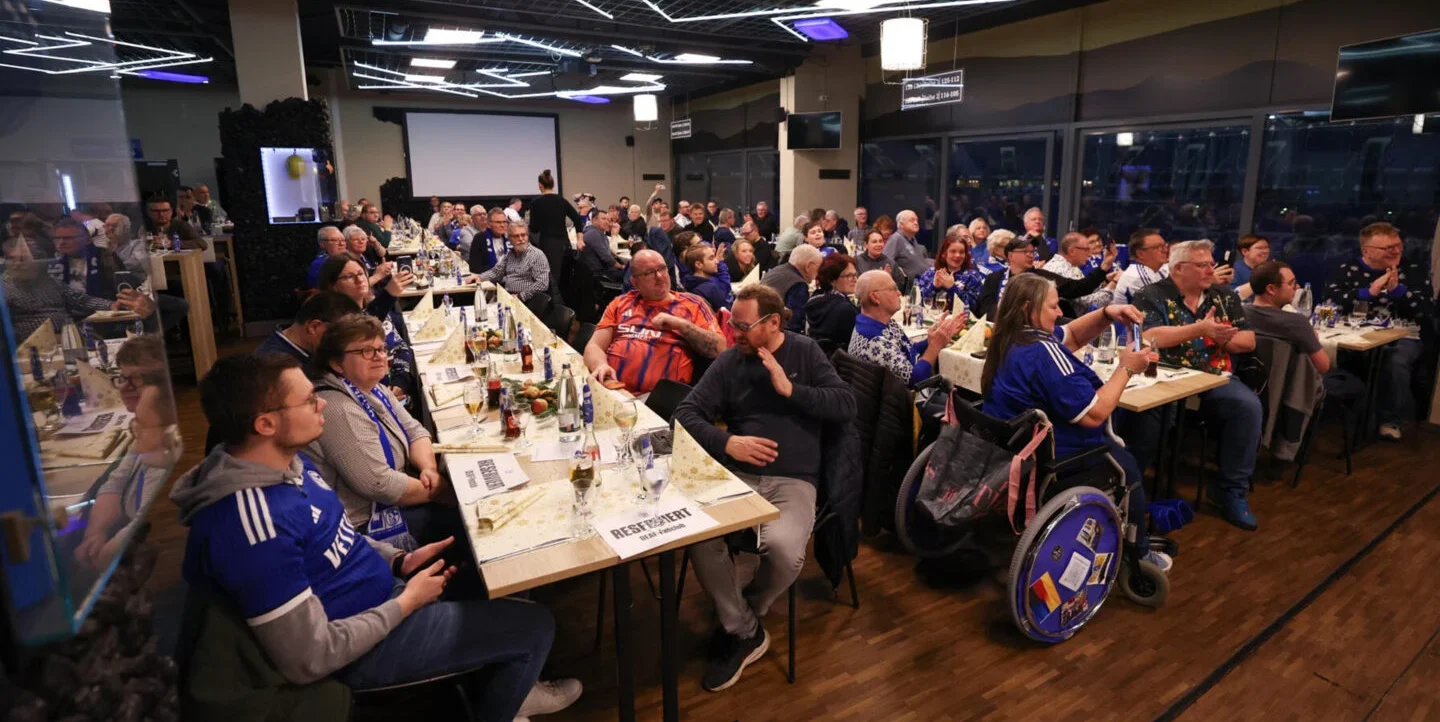 Eine Gruppe von Menschen sitzt an Tischen in einem gut beleuchteten Raum und nimmt an einer Veranstaltung teil. Die Teilnehmer tragen verschiedene Outfits, darunter auch Trikots, und einige sind in Rollstühlen. Die Tische sind mit Gläsern, Flaschen und Papieren gedeckt. Im Hintergrund ist eine Projektionsfläche zu sehen, und die Atmosphäre wirkt gesellig und aktiv.