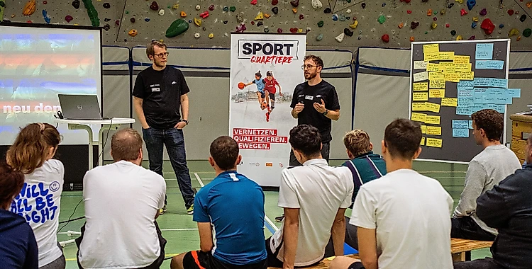 Zwei Referenten stehen vor einer Gruppe von Zuhörern in einem Raum mit Kletterwand. Die Referenten präsentieren eine Folie mit dem Titel "SPORT QUARTIERE" und den Untertiteln "VERNETZEN QUALIFIZIEREN WEITERGEHEN". Die Zuhörer sitzen auf Bänken und hören aufmerksam zu. Im Hintergrund sind bunte Klettergriffe an der Wand zu sehen.