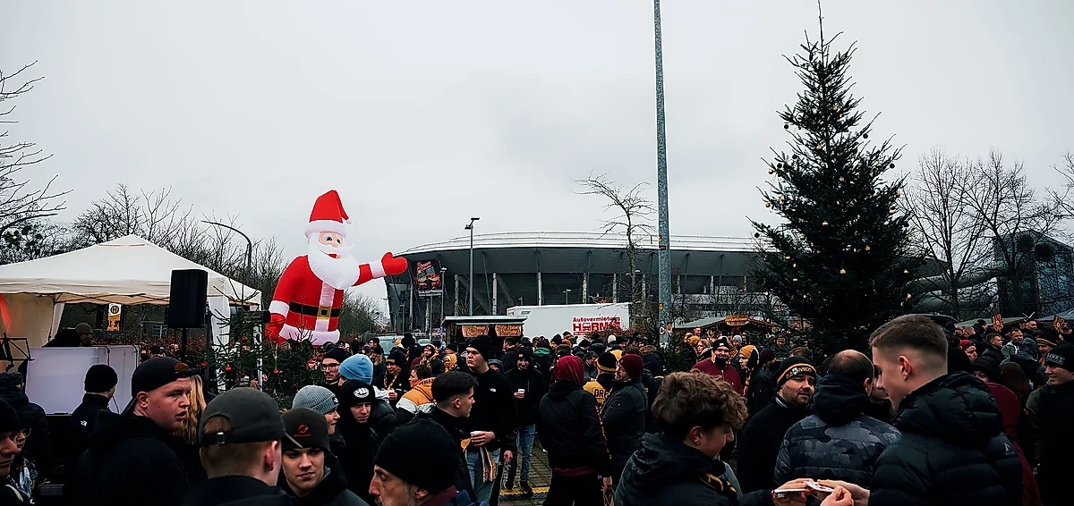 Eine große Menschenmenge versammelt sich auf einem Weihnachtsmarkt vor einem Stadion. Im Vordergrund steht eine große aufblasbare Weihnachtsmann-Figur, die mit einem Arm winkt. Die Besucher tragen warme Kleidung und scheinen sich an Ständen zu versammeln, die mit Weihnachtsdekoration geschmückt sind. Im Hintergrund ist ein großes Stadion zu sehen, das von kahlen Bäumen umgeben ist.