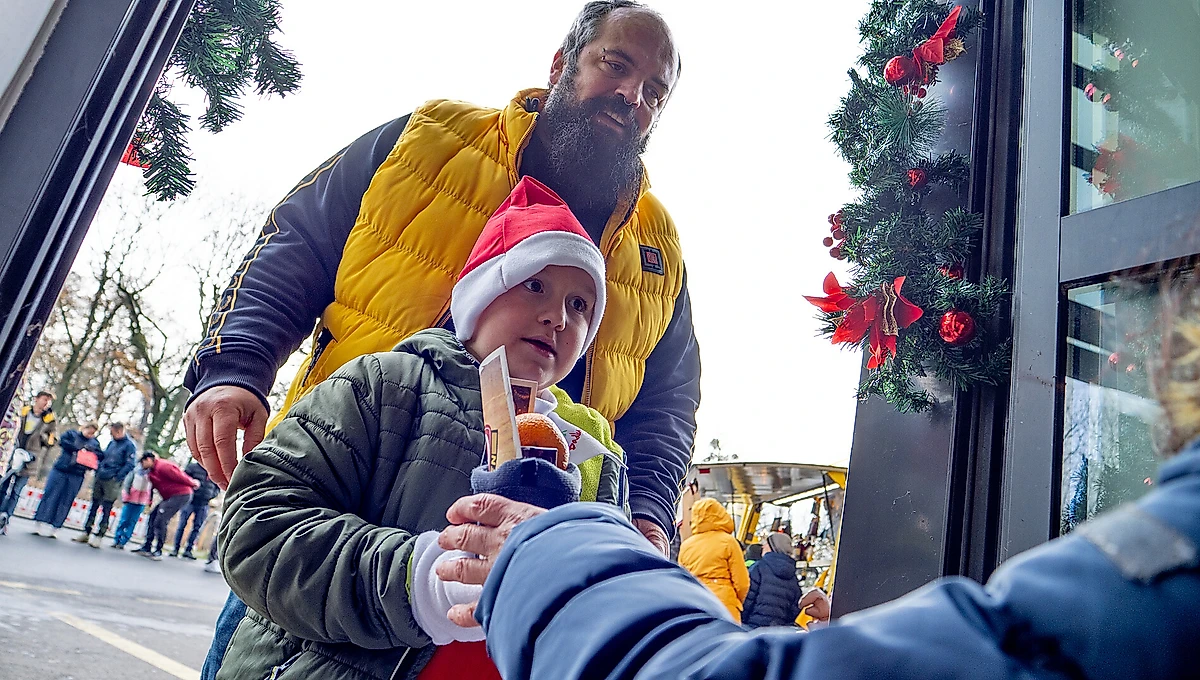 Ein Mann mit Bart und gelber Jacke trägt ein Kind mit roter Weihnachtsmütze auf dem Arm. Das Kind hält einen Lebkuchenmann in der Hand. Im Hintergrund sind weihnachtlich geschmückte Tannenbäume und Menschen in winterlicher Kleidung zu sehen.