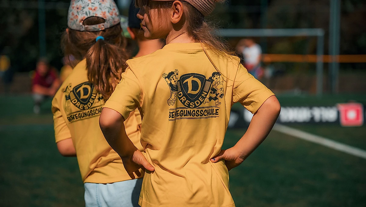 Zwei Kinder stehen auf einem Fußballfeld und tragen gelbe T-Shirts mit dem Aufdruck "Dresdner Bewegungsschule" und einem dazugehörigen Logo.