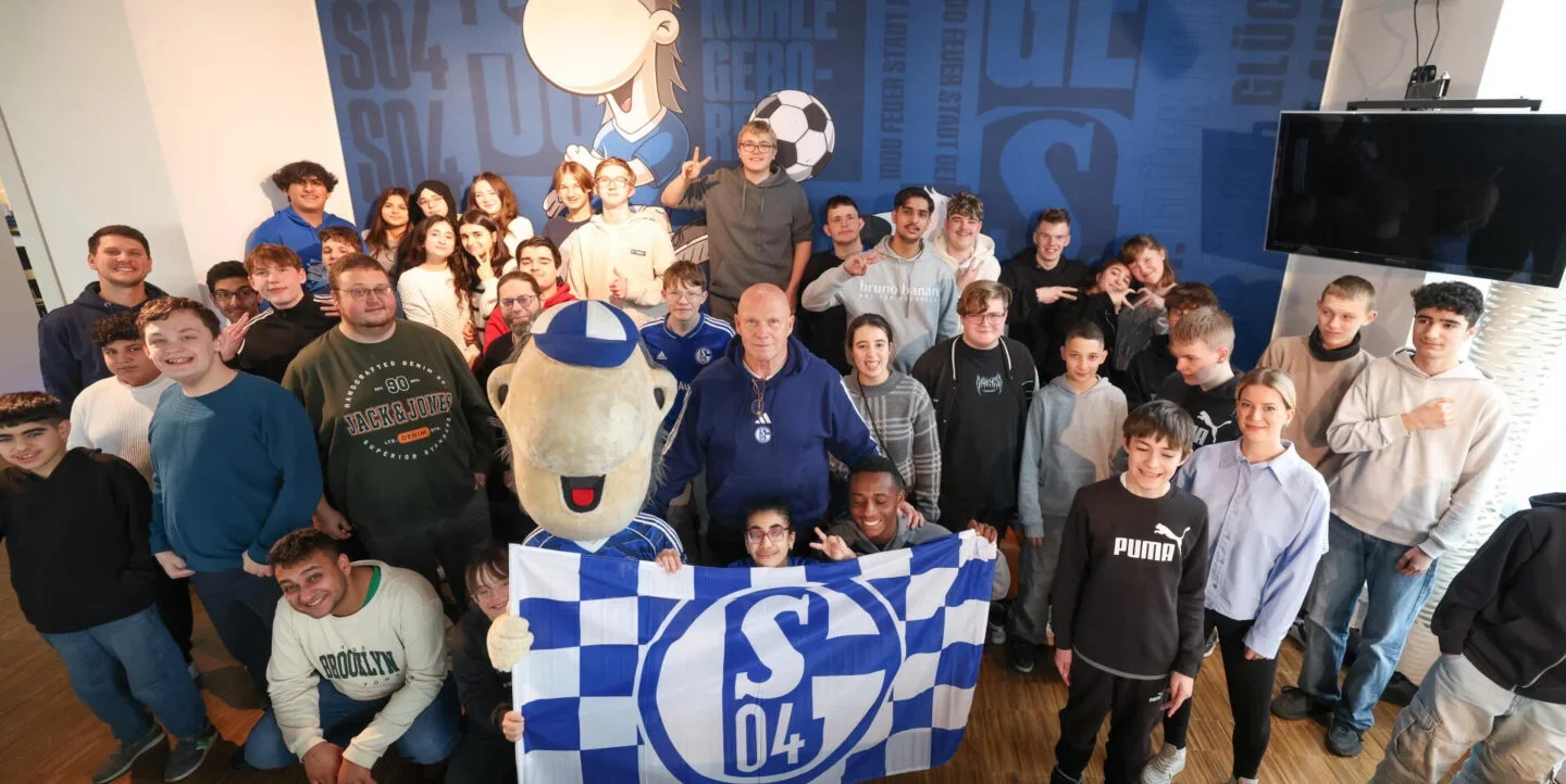 Eine Gruppe von Menschen, darunter Kinder, Jugendliche und Erwachsene, posieren für ein Gruppenfoto. Sie halten ein blau-weiß kariertes Banner mit dem Logo "S04", das für den Fußballverein Schalke 04 steht. Im Mittelpunkt des Fotos steht eine Person im Kostüm der Vereinsmaske von Schalke 04. Die Gruppe steht vor einer blau gestalteten Wand mit verschiedenen Aufschriften und Logos des Vereins. Die Atmosphäre wirkt fröhlich und gesellig, möglicherweise bei einem inklusiven Frühstücksereignis des Vereins.