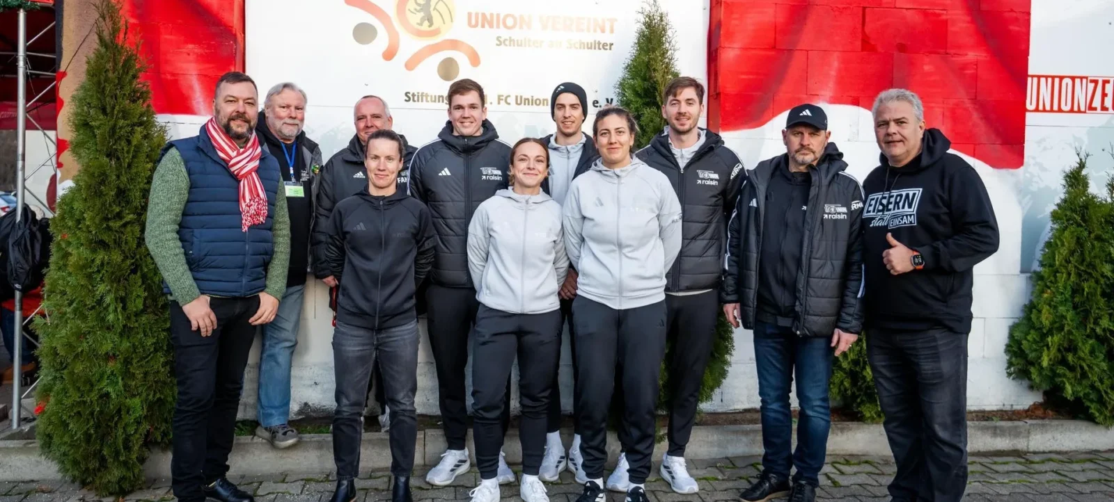 Eine Gruppe von Menschen steht vor einem Banner mit dem Logo und dem Namen "Union Vereint". Die Personen lächeln und posieren für ein Gruppenfoto. Sie tragen verschiedene Jacken und Kleidung, darunter einige mit dem Aufdruck "Eisern Union". Im Hintergrund sind rote und weiße Banner sowie grüne Pflanzen zu sehen.