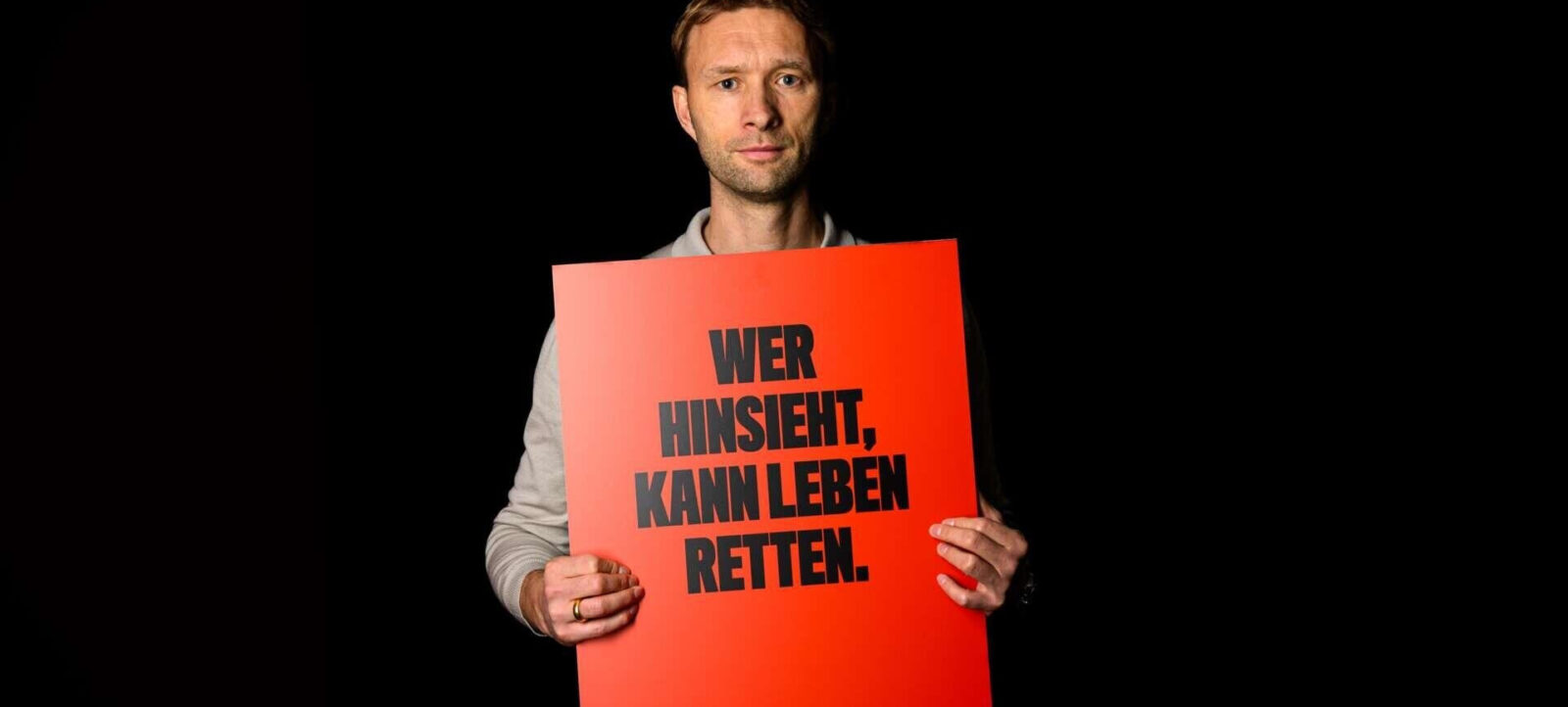 Ein Mann hält ein rotes Schild mit der Aufschrift "Wer hinsieht, kann Leben retten." Er steht vor einem schwarzen Hintergrund und blickt direkt in die Kamera.