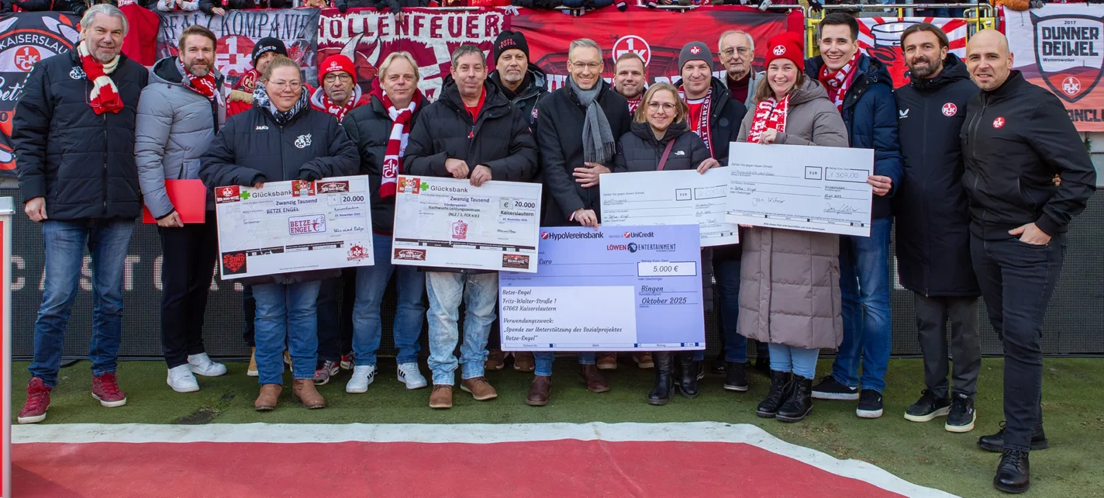 Eine Gruppe von Menschen steht auf einem Fußballfeld und hält Schecks in den Händen. Sie tragen warme Kleidung und einige von ihnen haben Schals mit den Farben und Logos des 1. FC Kaiserslautern. Im Hintergrund sind Fans mit Schals und Banner zu sehen. Die Schecks zeigen Spendenbeträge für soziale Projekte des Vereins.