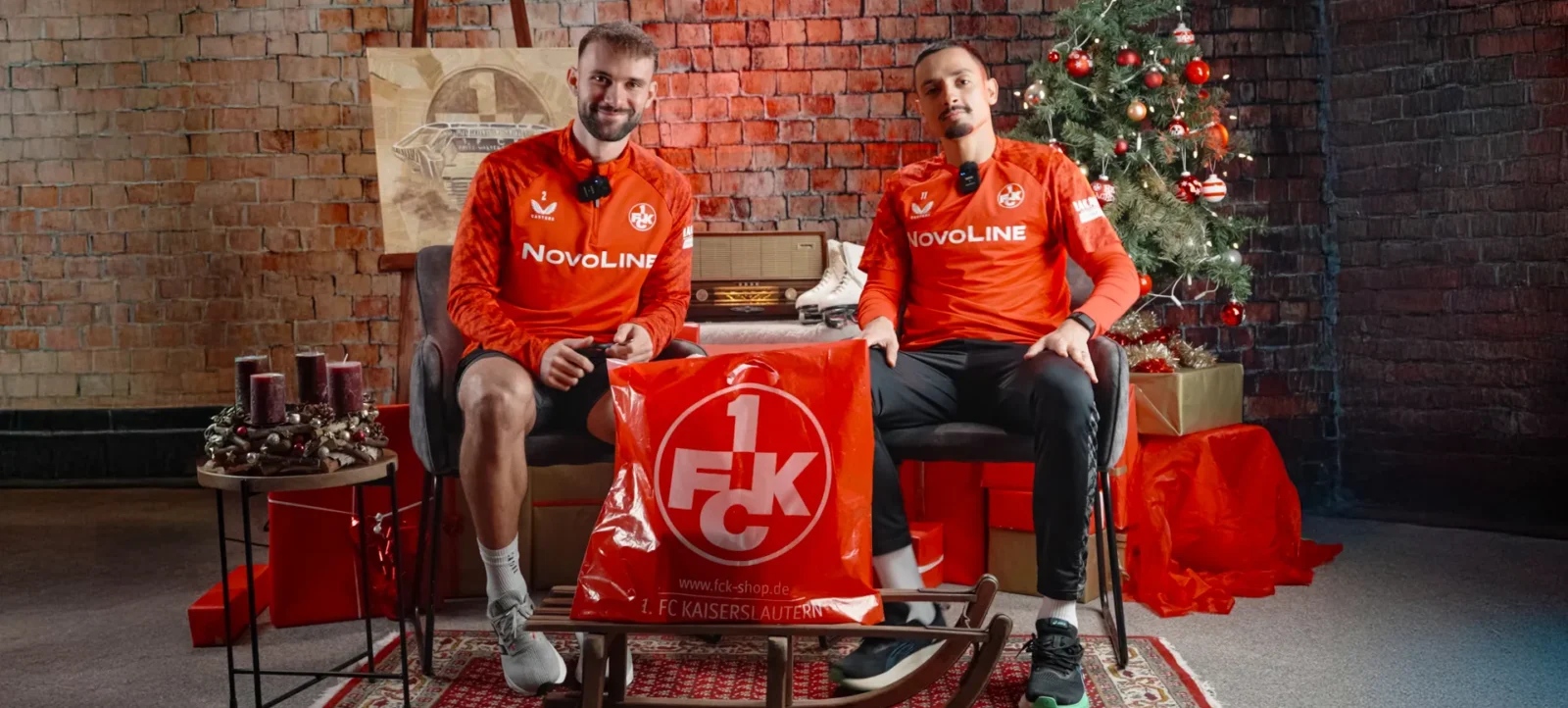 Zwei Fußballspieler des 1. FC Kaiserslautern in roten Trainingsjacken sitzen auf einem Schlitten vor einem weihnachtlich dekorierten Hintergrund. Sie halten einen roten Beutel mit dem Logo des 1. FC Kaiserslautern.