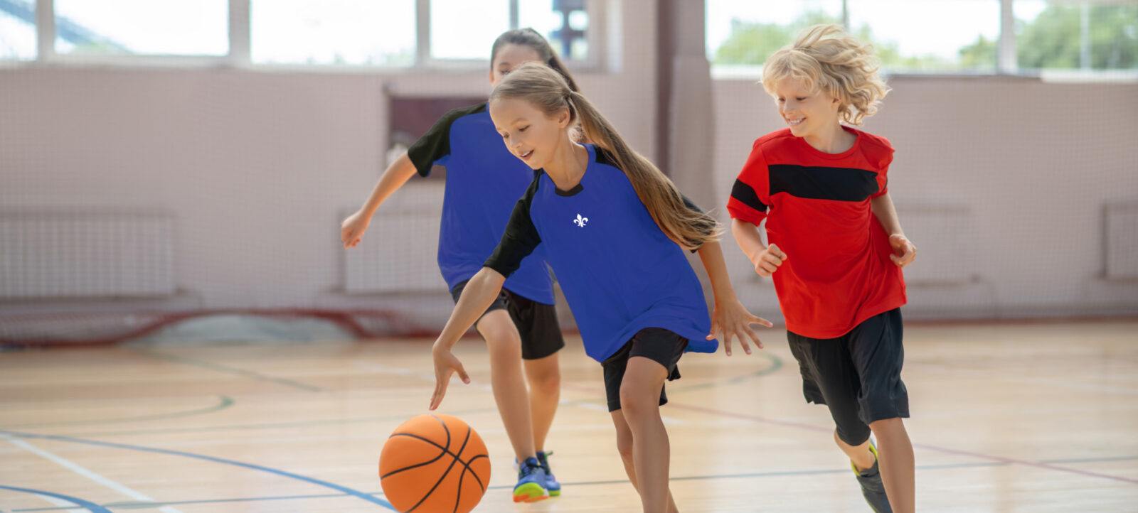 Drei Kinder spielen in einer Sporthalle Basketball. Ein Mädchen in blauem Trikot dribbelt den Ball, während zwei andere Kinder, eines in rotem Trikot und eines ebenfalls in blauem Trikot, ihr folgen. Die Halle hat große Fenster und einen Holzfußboden mit blauen Linien.