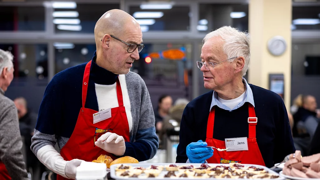 Zwei Männer in Schürzen stehen hinter einem Buffet und unterhalten sich. Sie tragen Namensschilder und bereiten Essen vor. Im Hintergrund sind weitere Personen und eine Küche zu sehen.