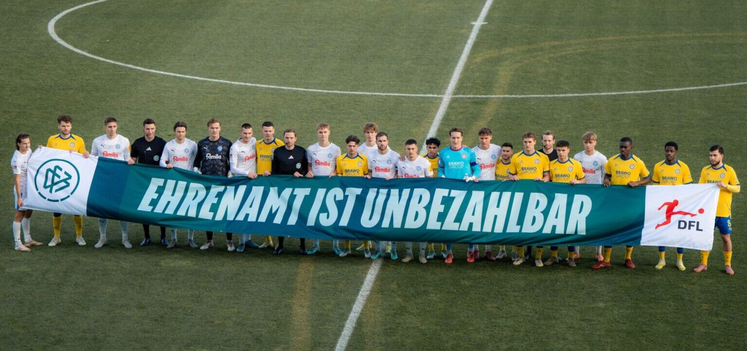 Eine Gruppe von Fußballspielern und Trainern steht auf dem Spielfeld und hält ein großes grünes Banner mit der Aufschrift "EHRENAMT IST UNBEZAHLBAR". Die Spieler tragen gelbe und weiße Trikots mit verschiedenen Sponsorenlogos. Links und rechts des Banners sind zwei weitere Banner mit den Logos der DFL und eines weiteren Sponsors zu sehen.