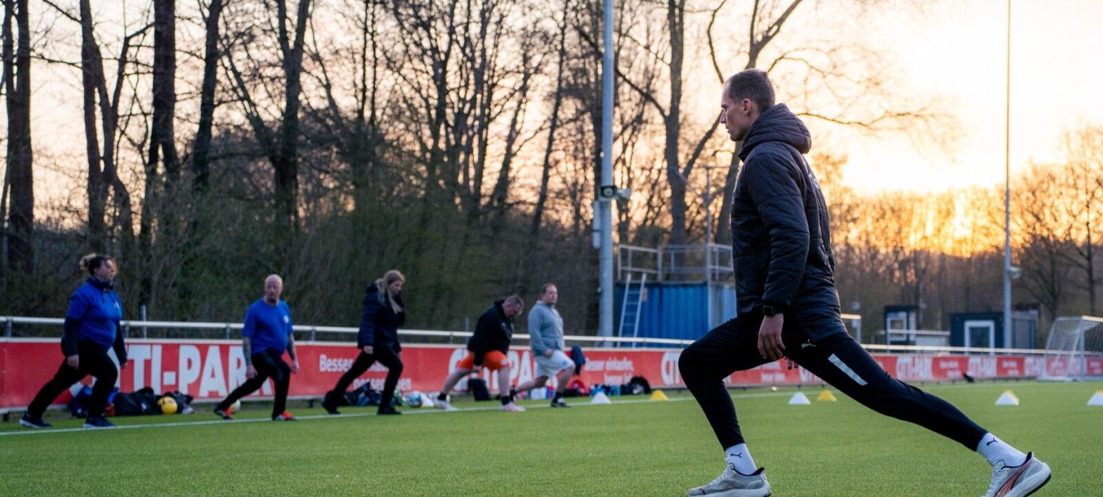 Ein Fußballer in schwarzer Trainingskleidung führt auf einem Rasenplatz ein Dehnungs- oder Aufwärmübung durch, während im Hintergrund weitere Personen ähnliche Übungen machen. Die Szene spielt bei Sonnenuntergang oder -aufgang, mit Bäumen und einer Werbebande im Hintergrund.