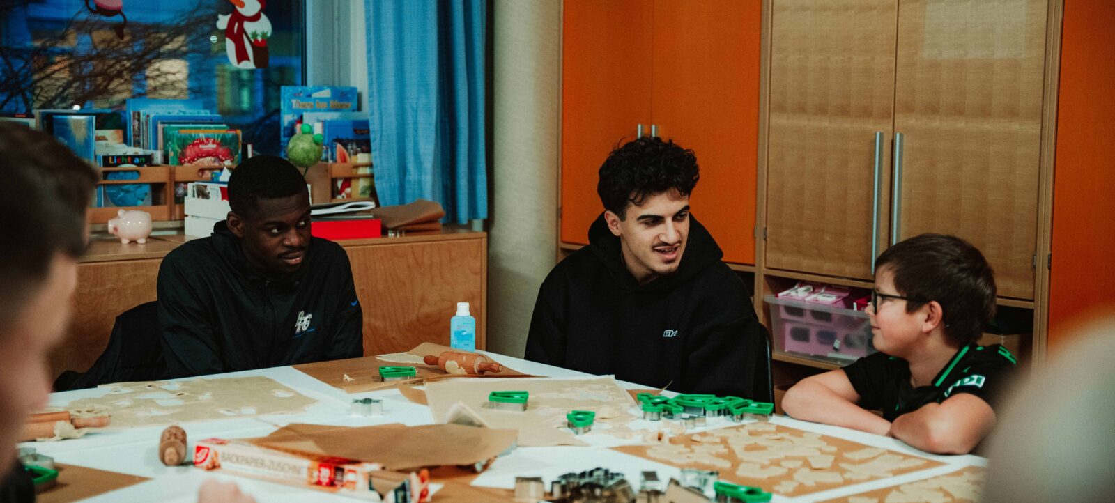 Zwei Personen sitzen an einem Tisch und unterhalten sich mit zwei Kindern, während sie gemeinsam an einem Bastelprojekt arbeiten. Der Tisch ist mit Bastelmaterialien wie Papier, Kleber und kleinen Figuren bedeckt. Im Hintergrund ist ein Regal mit Spielzeug und Dekorationen zu sehen, darunter ein Weihnachtsmann und ein Rentier.