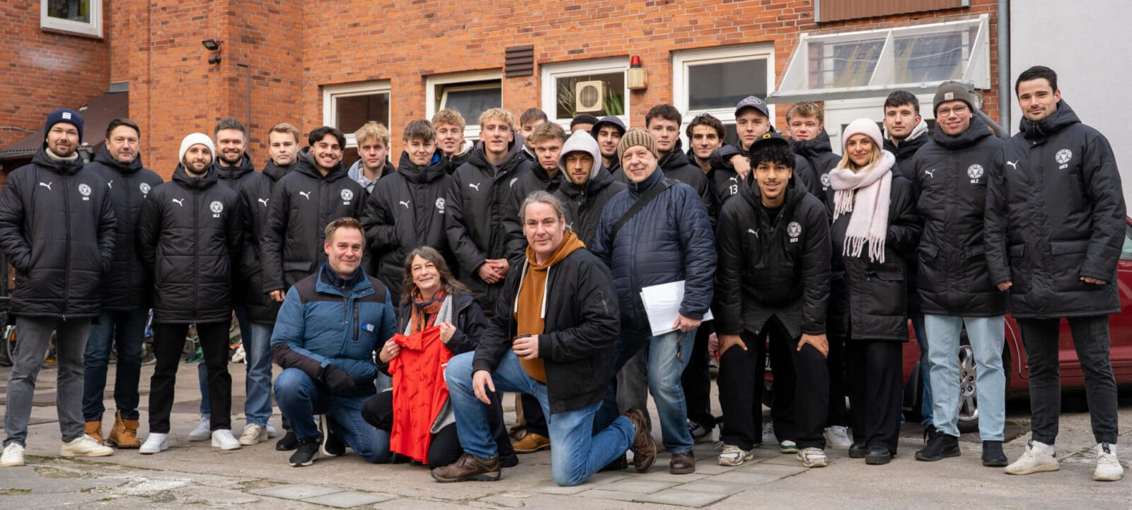 Eine Gruppe von Menschen, darunter Spieler und Betreuer eines Fußballteams, posieren für ein Gruppenfoto vor einem roten Backsteingebäude. Die meisten tragen schwarze Jacken mit dem Team-Logo, einige haben Mützen oder Schals an. Drei Personen in der vorderen Reihe knien, darunter eine Person, die ein rotes Tuch hält. Die Atmosphäre wirkt freundlich und gesellig.
