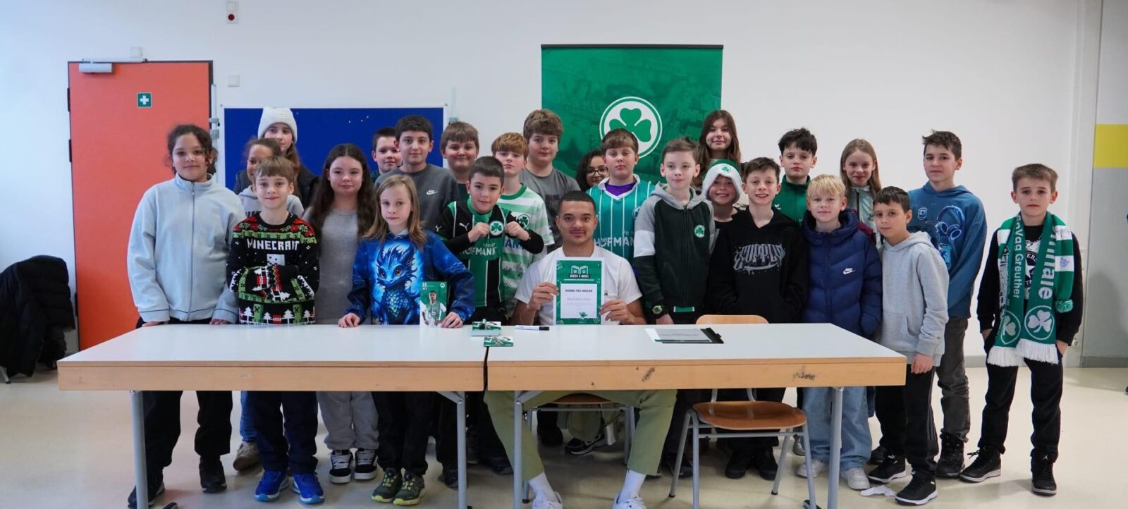 Gruppenfoto von Kindern und einem Erwachsenen in einem Klassenzimmer. Die Kinder tragen verschiedene T-Shirts und Jacken, einige mit Logos und Aufdrucken. Der Erwachsene sitzt an einem Tisch und hält ein grünes Schild mit einem Logo und Text. Im Hintergrund sind eine rote Tür und eine grüne Tafel mit einem dreiblättrigen Kleeblatt-Logo zu sehen.