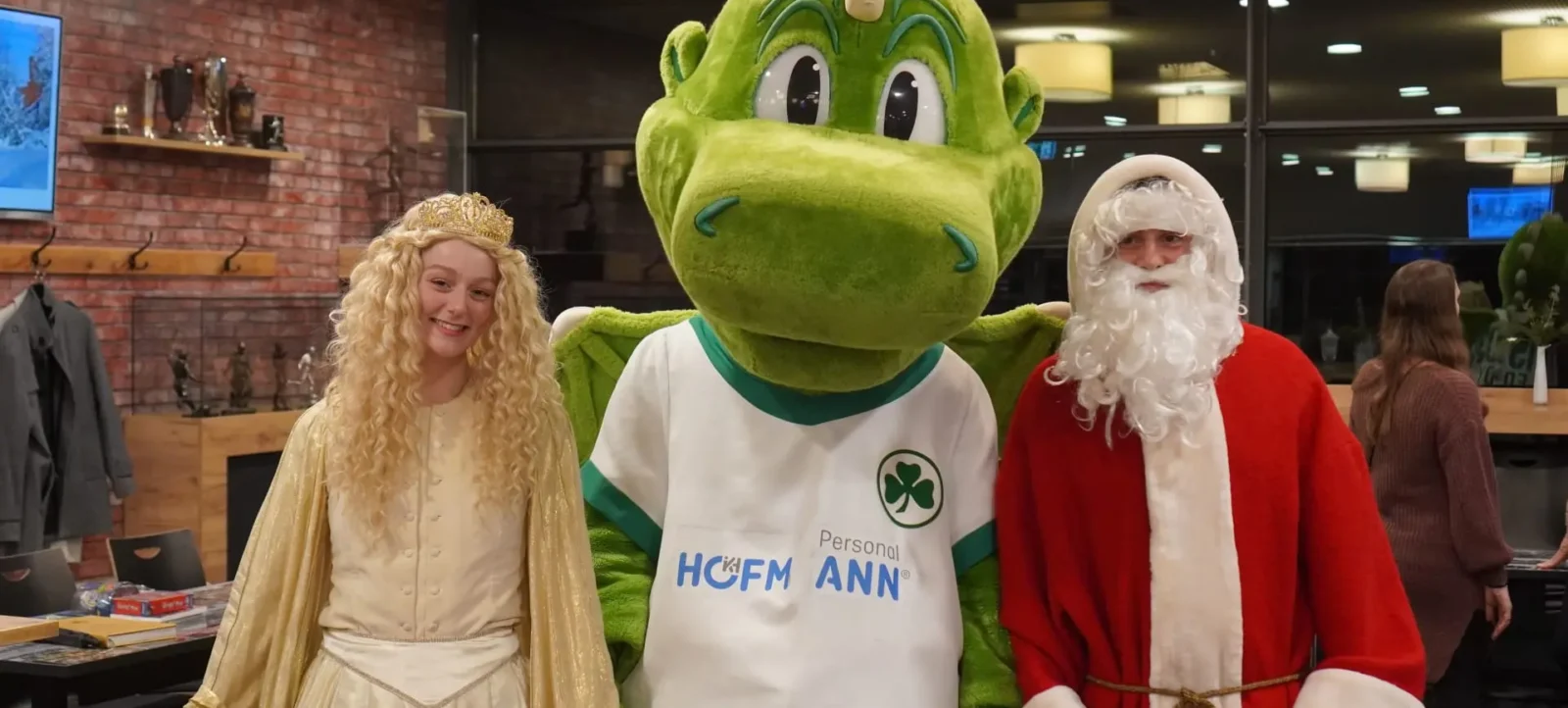 Drei Personen in Kostümen stehen nebeneinander: eine Person als Engel mit goldener Krone und goldenem Umhang, eine Person als Weihnachtsmann mit rotem Mantel und weißem Bart und eine Person als grüne Eidechse mit einem T-Shirt, das "Personal HOFMANN ANN" und ein Kleeblatt-Logo zeigt. Sie stehen in einem Raum mit Backsteinwand und Regalen im Hintergrund.