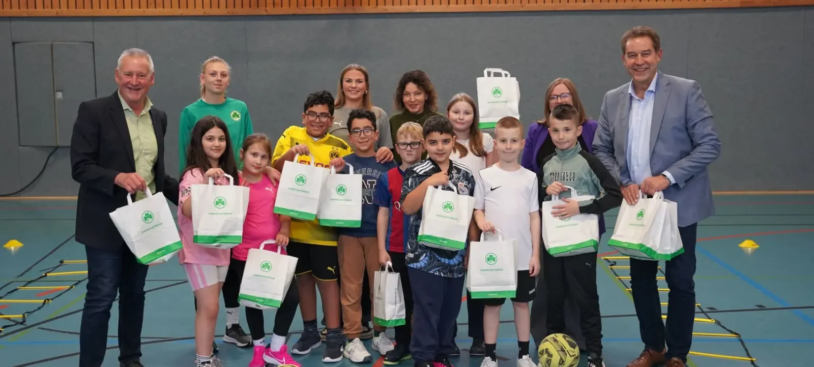 Gruppe von Kindern und Erwachsenen in einer Sporthalle, die Tüten mit dem Logo "Bundesliga Nachhaltigkeit" halten. Die Kinder und Erwachsenen lächeln in die Kamera. Im Hintergrund ist ein Basketballkorb zu sehen.