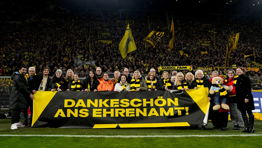 Eine Gruppe von Ehrenamtlichen steht auf dem Spielfeld eines Fußballstadions und hält ein großes Transparent mit der Aufschrift "Dankeschön ans Ehrenamt!". Im Hintergrund sind zahlreiche Fans zu sehen, die gelb-schwarze Schals und Fahnen schwenken. Die Szene ist Teil einer Veranstaltung, bei der die Arbeit der Ehrenamtlichen gewürdigt wird.