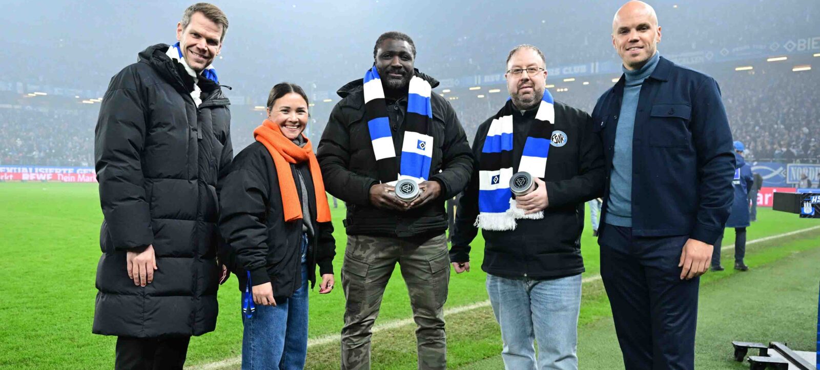 Gruppenfoto von fünf Personen auf einem Fußballfeld. Von links nach rechts: Dr. Eric Huwer (Vorstand Finanzen, HSV), Janin Scheer (Vorsitz, Kommission Engagement & Entwicklung, Hamburger Fußball-Verband), Babou Ceesay (Co- und Torwarttrainer 3. B-Jugend HSV), Dennis Bratschek (Fußball Jugend, FSV Harburg-Roenneburg e.V.), und Präsident Henrik Koencke (HSV e.V.). Alle tragen warme Kleidung und lächeln in die Kamera. Im Hintergrund sind Zuschauer auf den Tribünen zu sehen.