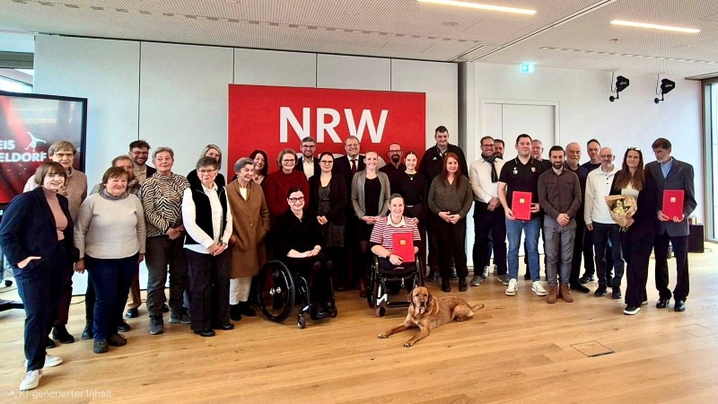 Gruppenfoto von Preisträgern und Gästen bei der Verleihung des Ehrenamtspreises NRW. Die Teilnehmer stehen vor einer großen roten Wand mit der Aufschrift "NRW". Einige Personen halten Urkunden oder Blumen in den Händen. Eine Person im Rollstuhl und ein Hund sind ebenfalls im Bild zu sehen.