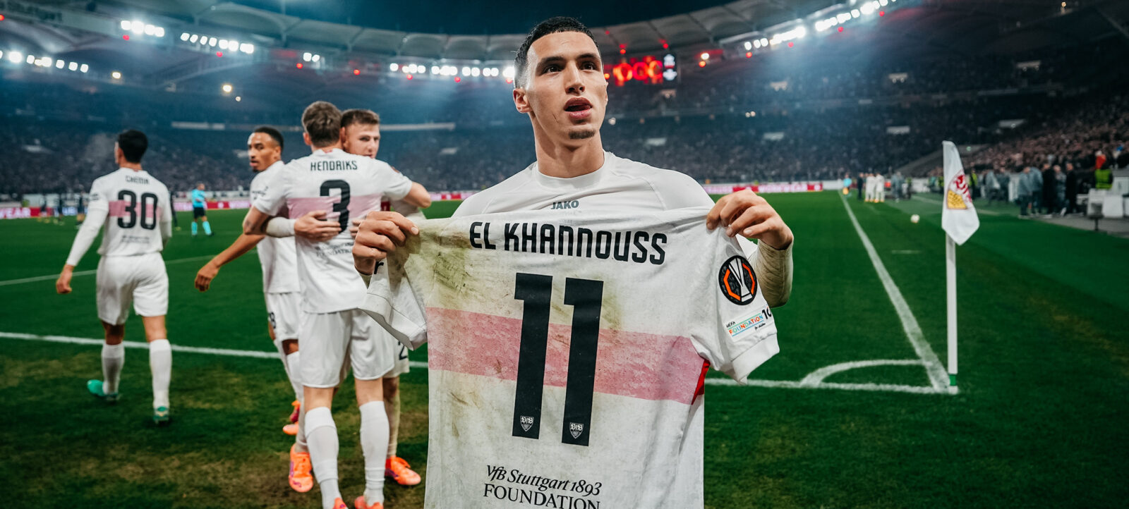 Ein Fußballspieler hält sein Trikot hoch, auf dem "EL KHANNOUSS" und die Nummer "11" zu sehen sind. Im Hintergrund sind weitere Spieler und ein volles Stadion zu erkennen.