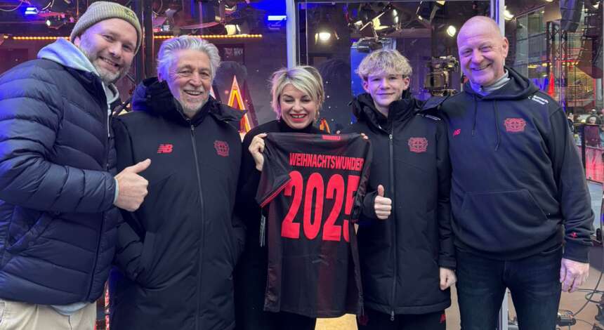 Eine Gruppe von fünf Personen steht zusammen und lächelt in die Kamera. Eine Person in der Mitte hält ein schwarzes T-Shirt mit der Aufschrift "Weihnachtswunder 2025" hoch. Alle tragen dunkle Jacken mit einem roten Logo auf der Brust. Der Hintergrund zeigt eine beleuchtete Weihnachtsdekoration.